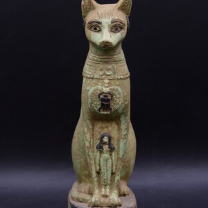 May include: A green and brown ceramic statue of a seated cat with a human figure on its chest. The cat has a scarab beetle on its chest and hieroglyphics on its base.