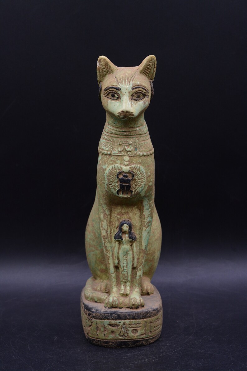 May include: A green and brown ceramic statue of a seated cat with a human figure on its chest. The cat has a scarab beetle on its chest and hieroglyphics on its base.