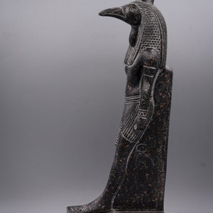 Puede incluir: Estatua de granito negro del dios egipcio Horus, representado como un hombre con cabeza de halc&oacute;n con una corona. La estatua est&aacute; de pie sobre una base rectangular con jerogl&iacute;ficos.