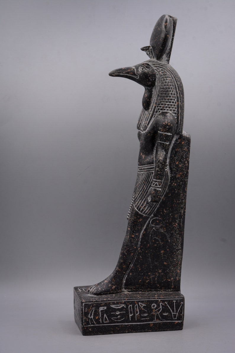 Puede incluir: Estatua de granito negro del dios egipcio Horus, representado como un hombre con cabeza de halc&oacute;n con una corona. La estatua est&aacute; de pie sobre una base rectangular con jerogl&iacute;ficos.