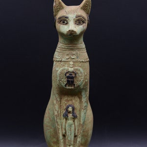 May include: A green and black Egyptian cat statue with a human figure carved into its chest. The statue is sitting on a base with hieroglyphics.