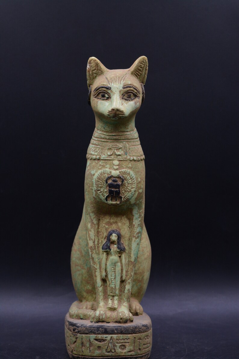 May include: A green and black Egyptian cat statue with a human figure carved into its chest. The statue is sitting on a base with hieroglyphics.