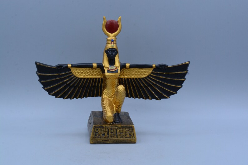 May include: A gold and black figurine of the Egyptian goddess Bastet, with outstretched wings and a red gem on her headdress. She is kneeling on a base with hieroglyphics.
