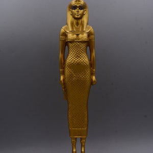 May include: A gold-coloured statue of a woman in a long dress, standing on a rectangular base. The statue has a detailed headdress and is posed with arms at her sides. The figure's dress has a scale-like pattern. The statue is approximately 30 cm tall.