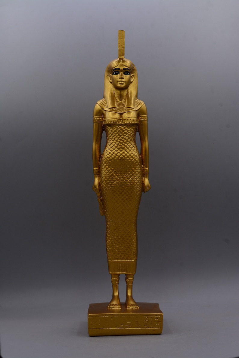 May include: A gold-coloured statue of a woman in a long dress, standing on a rectangular base. The statue has a detailed headdress and is posed with arms at her sides. The figure's dress has a scale-like pattern. The statue is approximately 30 cm tall.