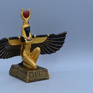 May include: A gold and black winged statue of the Egyptian goddess Isis, kneeling on a base with hieroglyphics. The statue has a red gem on its head.