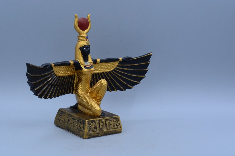 May include: A gold and black winged statue of the Egyptian goddess Isis, kneeling on a base with hieroglyphics. The statue has a red gem on its head.