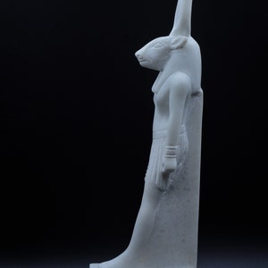 May include: White marble statue of an Egyptian goddess with a jackal head. The statue is standing on a rectangular base with hieroglyphics.