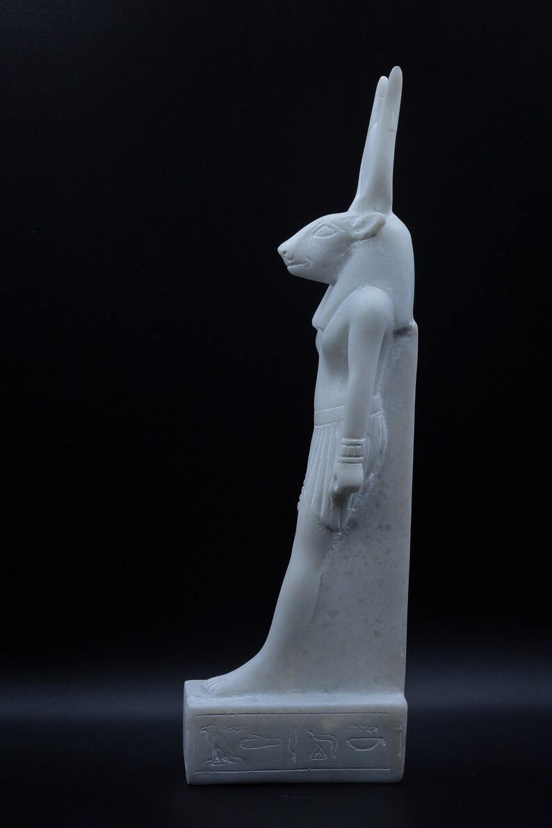 May include: White marble statue of an Egyptian goddess with a jackal head. The statue is standing on a rectangular base with hieroglyphics.