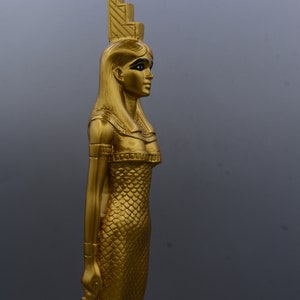 May include: A gold-coloured statue of an Egyptian figure, possibly a goddess or pharaoh, with a detailed headdress and scaled dress. The statue is in profile, holding an ankh symbol. The background is a solid grey.