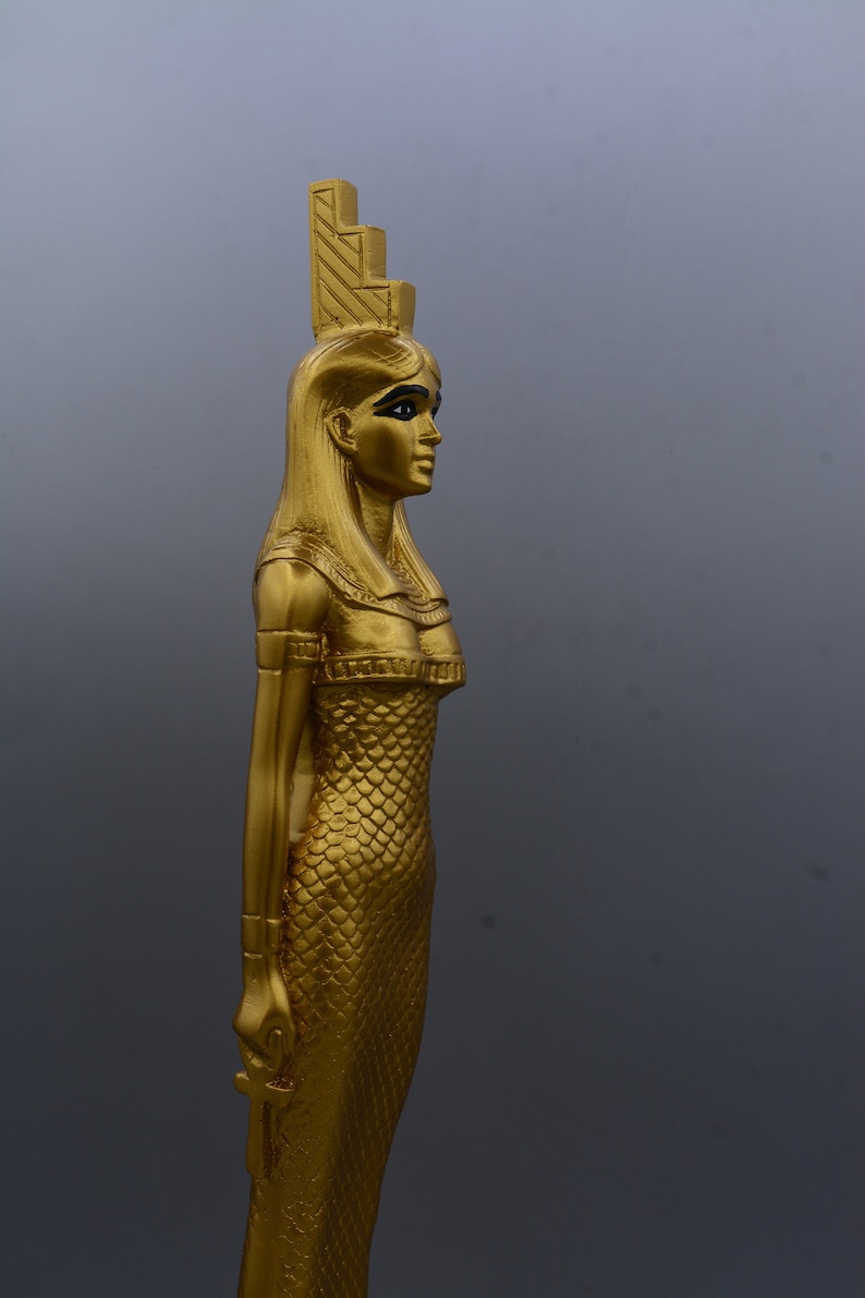 May include: A gold-coloured statue of an Egyptian figure, possibly a goddess or pharaoh, with a detailed headdress and scaled dress. The statue is in profile, holding an ankh symbol. The background is a solid grey.