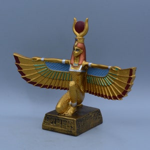 May include: Gold statue of the Egyptian goddess Isis with outstretched wings, wearing a red and gold crown and a blue and gold breastplate. The statue is standing on a base with hieroglyphics.