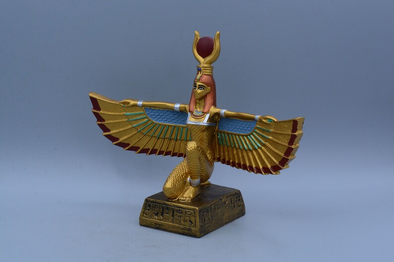 May include: Gold statue of the Egyptian goddess Isis with outstretched wings, wearing a red and gold crown and a blue and gold breastplate. The statue is standing on a base with hieroglyphics.