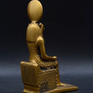 Peut inclure: Statue dor&eacute;e d'une figure assise, peut-&ecirc;tre une divinit&eacute;, avec une coiffe &eacute;labor&eacute;e et des v&ecirc;tements d&eacute;taill&eacute;s. La statue est sur une base rectangulaire et a une finition m&eacute;tallique lisse. Le fond est sombre.