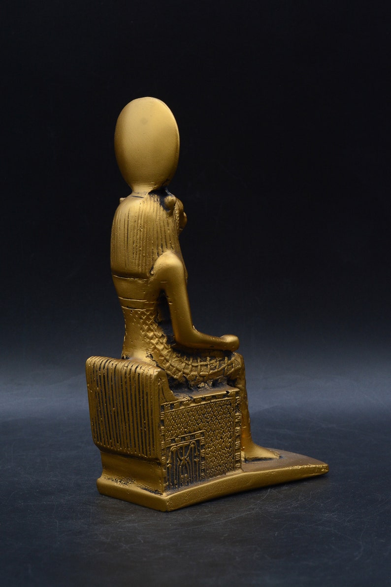 Peut inclure: Statue dor&eacute;e d'une figure assise, peut-&ecirc;tre une divinit&eacute;, avec une coiffe &eacute;labor&eacute;e et des v&ecirc;tements d&eacute;taill&eacute;s. La statue est sur une base rectangulaire et a une finition m&eacute;tallique lisse. Le fond est sombre.