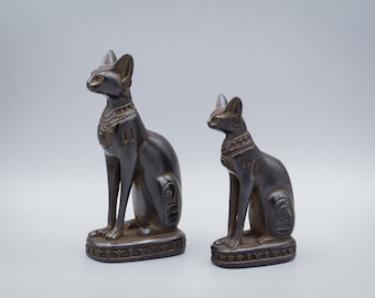 May include: Two black ceramic cat statues with intricate details. The larger statue is on the left and the smaller statue is on the right. Both statues are sitting with their heads turned to the left.