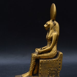 Peut inclure: Statue dor&eacute;e repr&eacute;sentant une figure assise avec une t&ecirc;te de lion et une haute coiffe allong&eacute;e. La statue est orn&eacute;e de motifs complexes et repose sur une base rectangulaire. Le fond est noir uni.