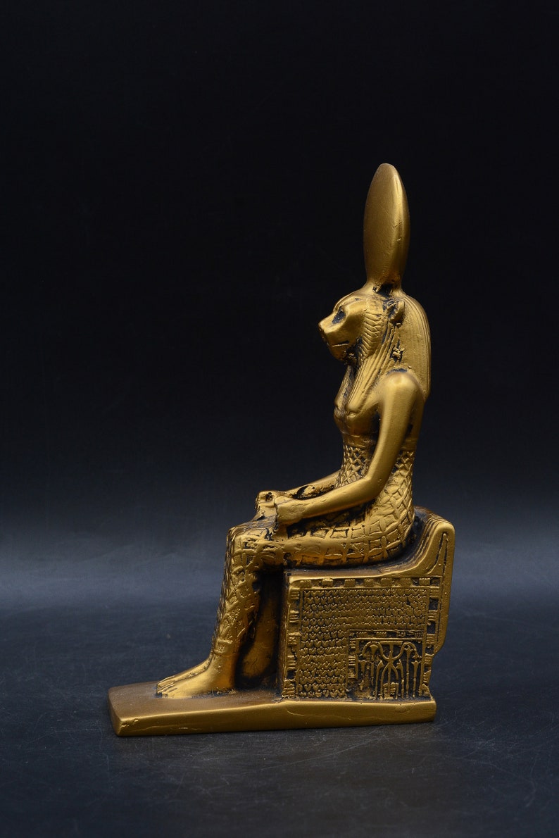 Peut inclure: Statue dor&eacute;e repr&eacute;sentant une figure assise avec une t&ecirc;te de lion et une haute coiffe allong&eacute;e. La statue est orn&eacute;e de motifs complexes et repose sur une base rectangulaire. Le fond est noir uni.