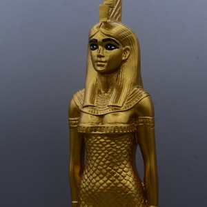 May include: A gold-coloured statue of a woman, possibly an Egyptian deity, with a detailed headdress and a scaled dress. The statue has black eyeliner and is set against a grey background. The figure is approximately 30 cm tall.