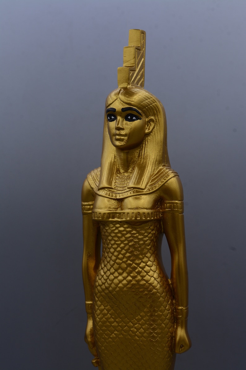 May include: A gold-coloured statue of a woman, possibly an Egyptian deity, with a detailed headdress and a scaled dress. The statue has black eyeliner and is set against a grey background. The figure is approximately 30 cm tall.