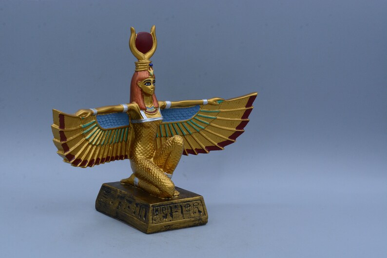 May include: A gold statue of the Egyptian goddess Isis with outstretched wings. She is kneeling on a base with hieroglyphics. The statue is painted with gold, red, and blue accents.