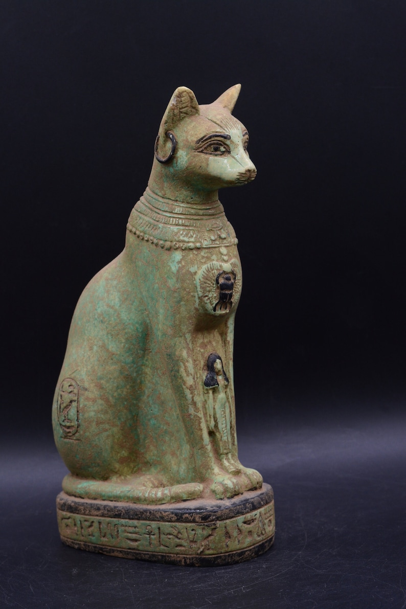 May include: A green and black Egyptian cat statue with hieroglyphics on the base. The cat is sitting with its head turned to the right. The statue is made of a hard material and has a smooth finish.