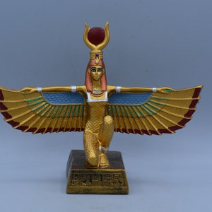 May include: Gold statue of the Egyptian goddess Isis with outstretched wings, wearing a crown with a red sun disc, and standing on a gold base with hieroglyphs.