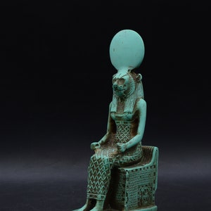 Peut inclure: Statue turquoise repr&eacute;sentant une figure assise &agrave; t&ecirc;te de lion, v&ecirc;tue d'une robe &agrave; motifs et d'une coiffe avec un disque solaire. La figure est assise sur une structure en forme de tr&ocirc;ne avec des d&eacute;tails hi&eacute;roglyphiques, sur fond noir.