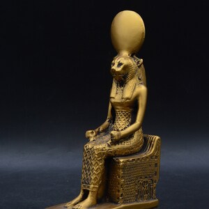 Peut inclure: Statue dor&eacute;e d'une figure assise &agrave; t&ecirc;te de lion, portant une coiffe et assise sur un tr&ocirc;ne d&eacute;taill&eacute;. La statue pr&eacute;sente des d&eacute;tails complexes et se d&eacute;tache sur un fond sombre.