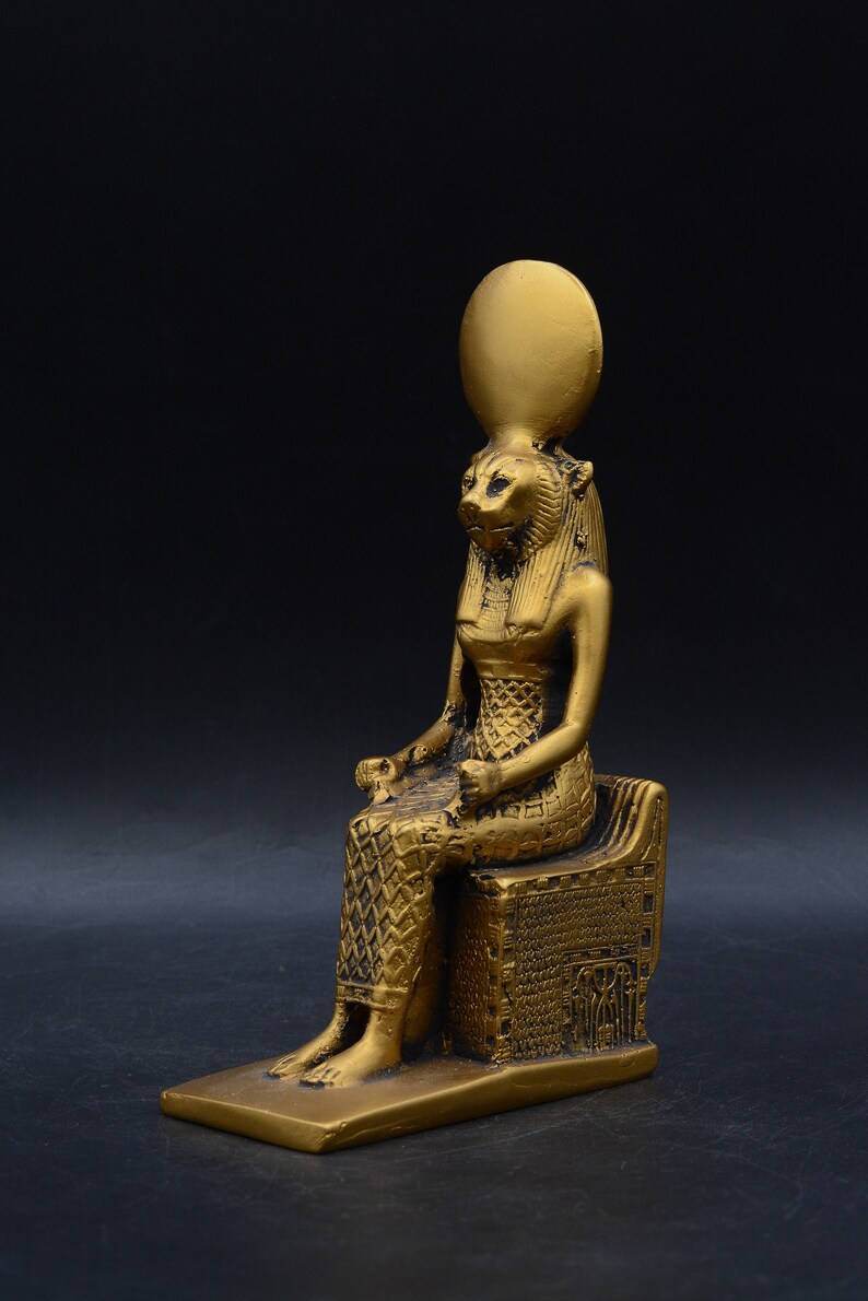 Peut inclure: Statue dor&eacute;e d'une figure assise &agrave; t&ecirc;te de lion, portant une coiffe et assise sur un tr&ocirc;ne d&eacute;taill&eacute;. La statue pr&eacute;sente des d&eacute;tails complexes et se d&eacute;tache sur un fond sombre.