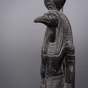 Puede incluir: Una estatua de granito negro del dios egipcio Horus, representado como un hombre con cabeza de halc&oacute;n con un disco solar en la cabeza. La estatua est&aacute; detallada con tallas intrincadas y una superficie texturizada.