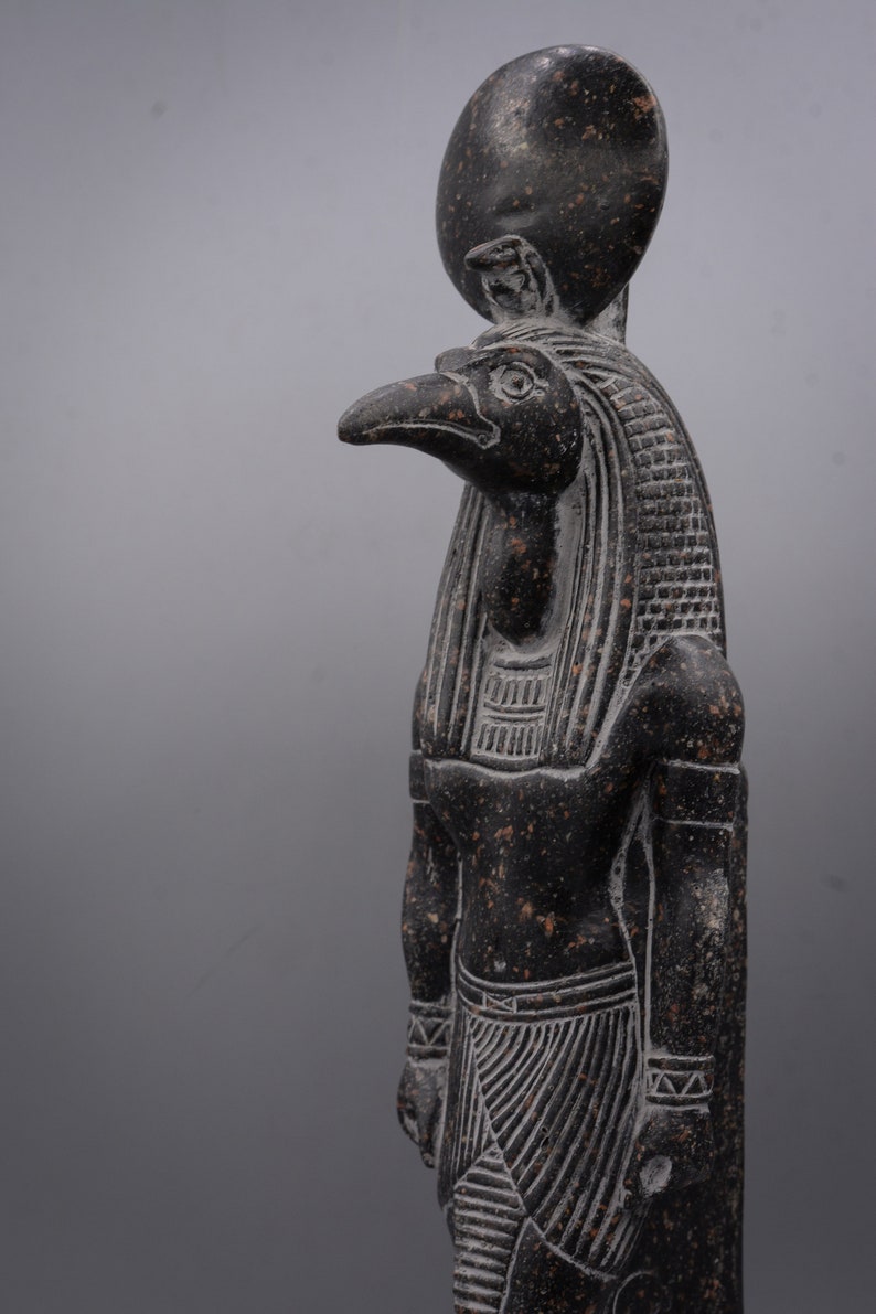 Puede incluir: Una estatua de granito negro del dios egipcio Horus, representado como un hombre con cabeza de halc&oacute;n con un disco solar en la cabeza. La estatua est&aacute; detallada con tallas intrincadas y una superficie texturizada.