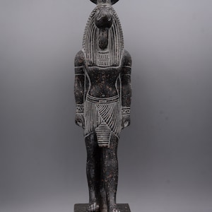 Puede incluir: Una estatua de granito negro del dios egipcio Anubis, representado como un humano con cabeza de chacal con un disco solar en la cabeza. La estatua est&aacute; de pie sobre una base rectangular con jerogl&iacute;ficos.