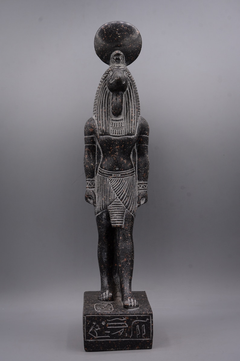 Puede incluir: Una estatua de granito negro del dios egipcio Anubis, representado como un humano con cabeza de chacal con un disco solar en la cabeza. La estatua est&aacute; de pie sobre una base rectangular con jerogl&iacute;ficos.