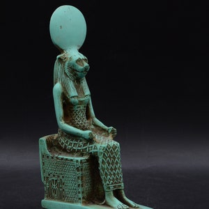 Peut inclure: Une statue turquoise d'une divinit&eacute; &eacute;gyptienne, peut-&ecirc;tre Bastet, assise sur un tr&ocirc;ne. La figure a une t&ecirc;te de f&eacute;lin, une coiffe &eacute;labor&eacute;e et des v&ecirc;tements &agrave; motifs d&eacute;taill&eacute;s. La statue est plac&eacute;e sur un fond noir.