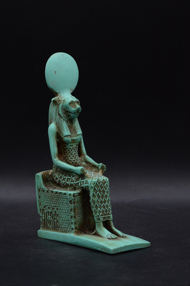 Peut inclure: Une statue turquoise d'une divinit&eacute; &eacute;gyptienne, peut-&ecirc;tre Bastet, assise sur un tr&ocirc;ne. La figure a une t&ecirc;te de f&eacute;lin, une coiffe &eacute;labor&eacute;e et des v&ecirc;tements &agrave; motifs d&eacute;taill&eacute;s. La statue est plac&eacute;e sur un fond noir.