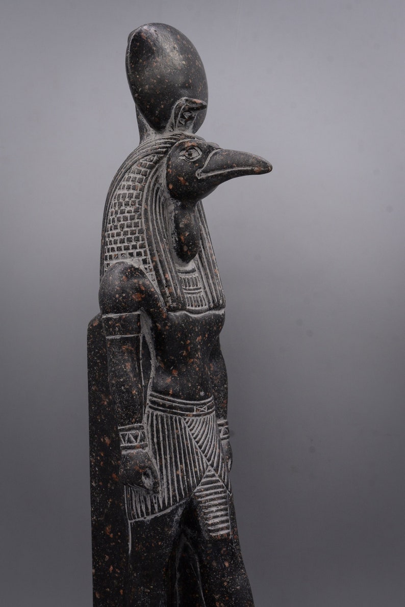 Puede incluir: Una estatua de piedra negra del dios egipcio Horus, representada como un hombre con cabeza de halc&oacute;n con un disco solar en la cabeza. La estatua est&aacute; detallada con intrincados tallados y una superficie texturizada.