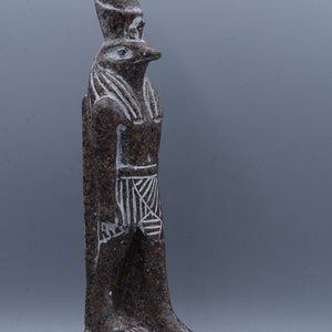 K&ouml;nnte beinhalten: Eine braune Granitstatue des &auml;gyptischen Gottes Horus, dargestellt als falkenk&ouml;pfiger Mann. Die Statue steht auf einem Sockel mit Hieroglyphen.