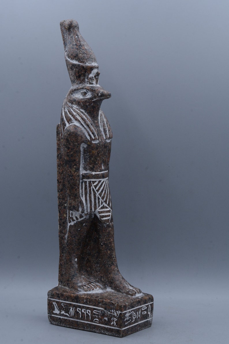 K&ouml;nnte beinhalten: Eine braune Granitstatue des &auml;gyptischen Gottes Horus, dargestellt als falkenk&ouml;pfiger Mann. Die Statue steht auf einem Sockel mit Hieroglyphen.