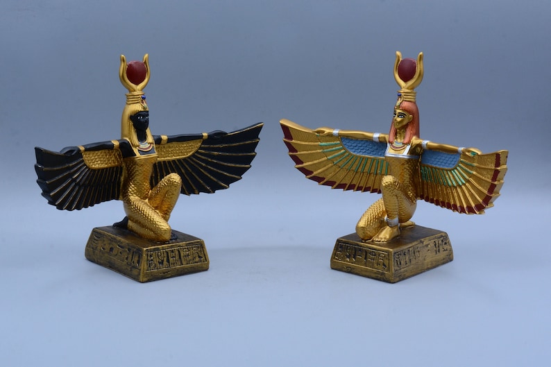 May include: Two gold-painted statues of the Egyptian goddess Isis with outstretched wings. One statue has black wings and the other has gold and blue wings.