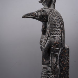 Puede incluir: Una estatua de granito negro del dios egipcio Horus, representado como un hombre con cabeza de halc&oacute;n y un alto tocado. La estatua est&aacute; tallada en un estilo detallado y realista, con intrincados detalles en las plumas, el tocado y la ropa.