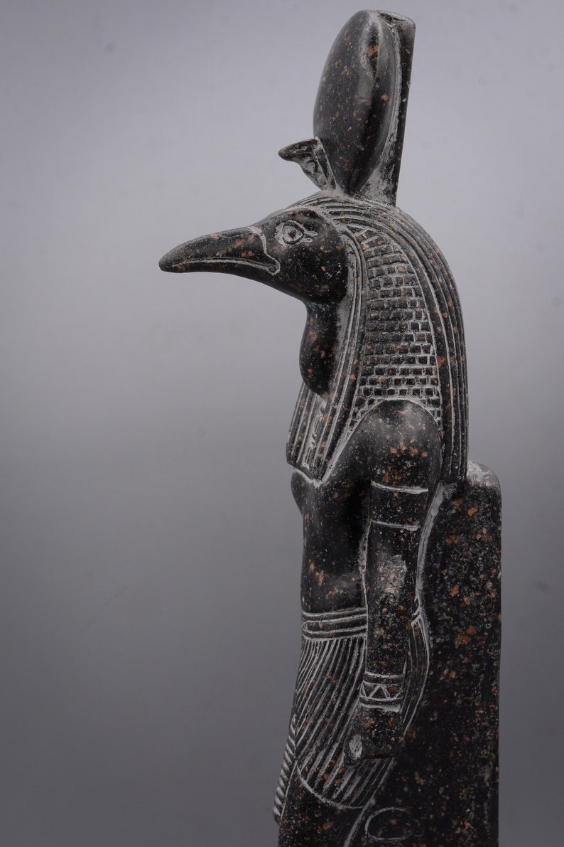 Puede incluir: Una estatua de granito negro del dios egipcio Horus, representado como un hombre con cabeza de halc&oacute;n y un alto tocado. La estatua est&aacute; tallada en un estilo detallado y realista, con intrincados detalles en las plumas, el tocado y la ropa.