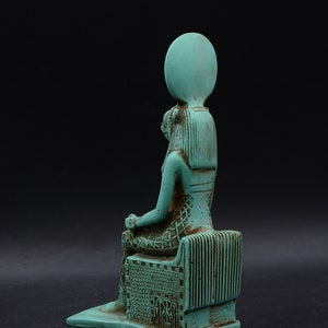 Peut inclure: Une statue turquoise d'une figure assise, possiblement une divinit&eacute;, avec une coiffe &eacute;labor&eacute;e et des sculptures d&eacute;taill&eacute;es. La figure est assise sur un tr&ocirc;ne avec des lignes verticales et des motifs complexes, sur fond sombre.