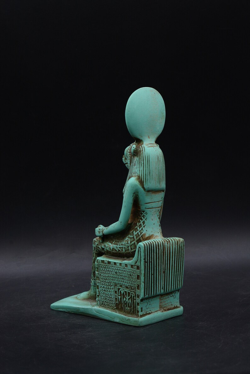 Peut inclure: Une statue turquoise d'une figure assise, possiblement une divinit&eacute;, avec une coiffe &eacute;labor&eacute;e et des sculptures d&eacute;taill&eacute;es. La figure est assise sur un tr&ocirc;ne avec des lignes verticales et des motifs complexes, sur fond sombre.