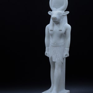 May include: White marble statue of an Egyptian god with a bull's head, wearing a headdress and a kilt. The statue is standing on a base with hieroglyphs.