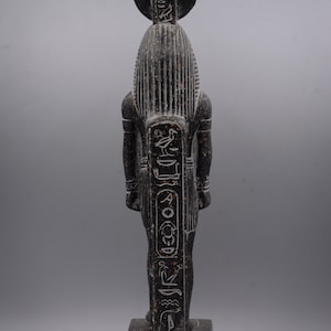 Puede incluir: Estatua de un dios egipcio de granito negro con jerogl&iacute;ficos en la parte delantera y trasera. La estatua est&aacute; de pie sobre una base cuadrada.