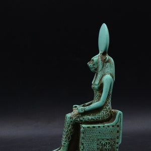 Peut inclure: Statue turquoise d'une figure assise &agrave; t&ecirc;te f&eacute;line, possiblement une divinit&eacute; &eacute;gyptienne. La figure est orn&eacute;e de d&eacute;tails complexes, dont une robe et une coiffe &agrave; motifs. La statue est pos&eacute;e sur un fond sombre.