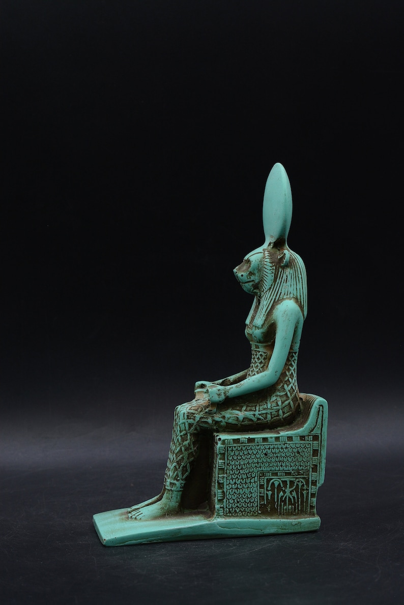 Peut inclure: Statue turquoise d'une figure assise &agrave; t&ecirc;te f&eacute;line, possiblement une divinit&eacute; &eacute;gyptienne. La figure est orn&eacute;e de d&eacute;tails complexes, dont une robe et une coiffe &agrave; motifs. La statue est pos&eacute;e sur un fond sombre.