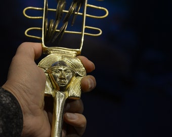May include: A gold-colored scepter with a female head at the top and a cage-like structure holding round objects. The scepter is held in a person's hand.