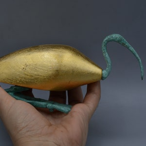 Peut inclure: Sculpture d'oiseau en feuille d'or avec un cou et des pattes verts. L'oiseau est en position accroupie, la tête tournée sur le côté.